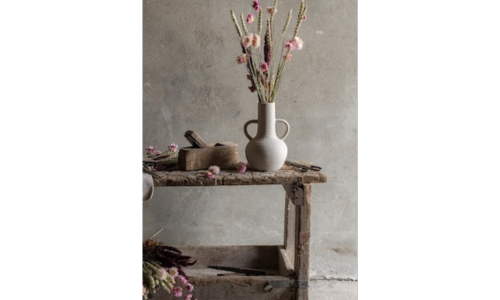 A rustic vase on a wooden bench with pink flowers arranged inside